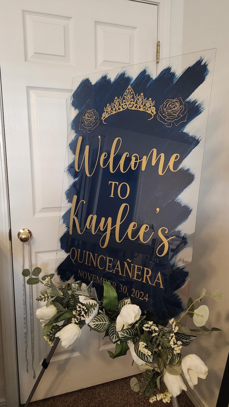 Op de afbeelding: Een blauw en gouden welkomsbord voor een Quincea&ntilde;era. Het bord zegt "Welkom bij Kaylee's Quincea&ntilde;era op 30 november 2024".