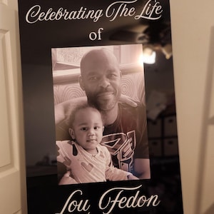 May include: A black framed memorial sign on a wooden easel. The sign reads "Celebrating The Life of Lou Fedon" in white script. The frame contains a black and white photo of a man and a child.