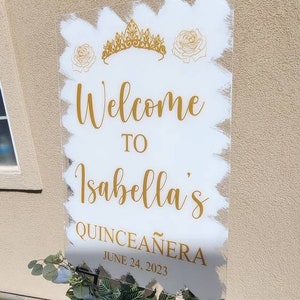 Op de afbeelding: Een wit acryl bord met gouden letters die "Welkom bij Isabella's Quincea&ntilde;era op 24 juni 2023" aankondigen. Het bord is versierd met een gouden kroon en witte rozen.
