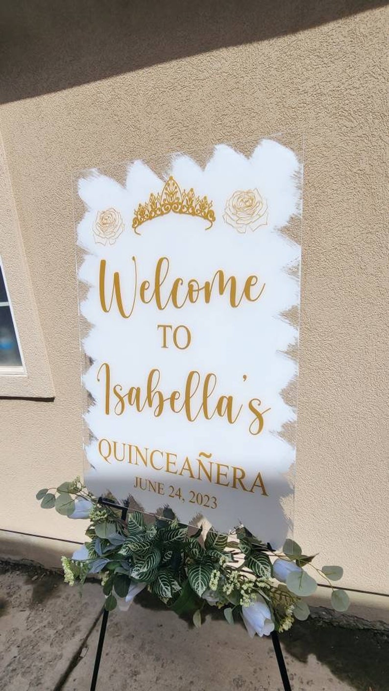 Op de afbeelding: Een wit acryl bord met gouden letters die "Welkom bij Isabella's Quincea&ntilde;era op 24 juni 2023" aankondigen. Het bord is versierd met een gouden kroon en witte rozen.