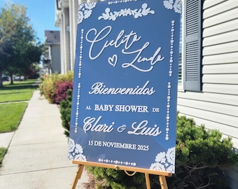 May include: A blue baby shower welcome sign on a gold easel. The sign has white floral accents and text that reads "Culito Linda Bienvenidos al Baby Shower de Clari & Luis 15 de Noviembre 2025."