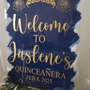 Op de afbeelding: Een blauw en gouden welkomsbord voor een Quincea&ntilde;era. Het bord zegt "Welkom bij Jaslene's Quincea&ntilde;era 8 februari 2025".