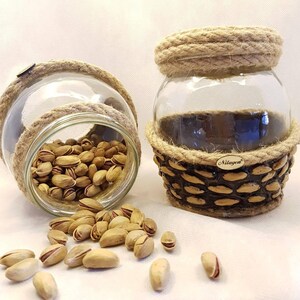 May include: Two glass jars with rope and woven brown accents, filled with pistachios. The jars are on a white surface with pistachios scattered around them. The label on the jar reads "Nilayeri".