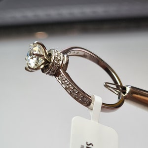 May include: A silver ring with a large, clear diamond set in a prong setting. The ring is surrounded by smaller diamonds. The ring is attached to a security tag that reads "SECURITY TAG ITEM NOT RETURNABLE IF REMOVED L.F.J."