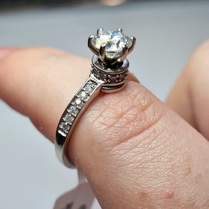 May include: A silver ring featuring a large, clear, round gemstone set in a crown-like design. The band is embellished with smaller, clear gemstones.