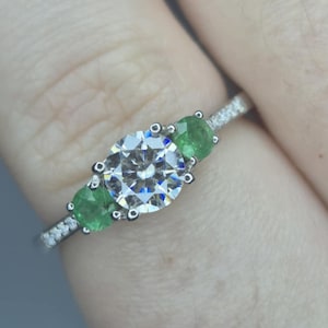 Puede incluir: Un anillo de plata con una piedra central grande, redonda y transparente, flanqueada por dos piedras verdes más pequeñas en forma de pera. La banda está incrustada con pequeñas piedras transparentes. El anillo está en un dedo.