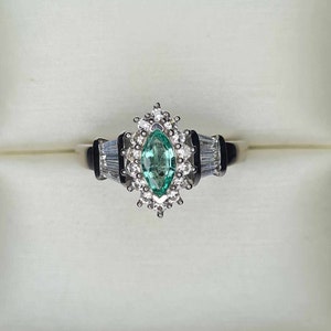 Puede incluir: Un anillo de plata con una esmeralda de forma marquesa rodeada de diamantes más pequeños. El anillo está engastado en una banda de metal blanco.