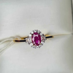 Puede incluir: Un anillo de oro con un gran rubí ovalado rodeado por un halo de diamantes más pequeños. El anillo tiene una banda simple.