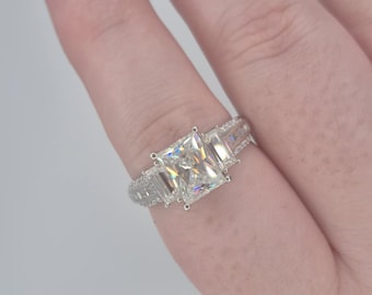 May include: A silver ring with a large, rectangular, clear gemstone surrounded by smaller, clear gemstones. The ring is on a finger.