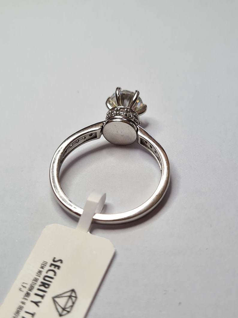 May include: A silver ring featuring a large clear cubic zirconia stone set in a crown-like design. The ring is displayed on a white background with a white tag that reads "SECURITY TAG ITEM NOT RETURNABLE IF REMOVED".