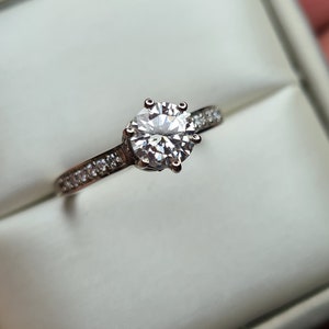 May include: A silver engagement ring with a large, round, clear diamond centre stone and a band of smaller diamonds.