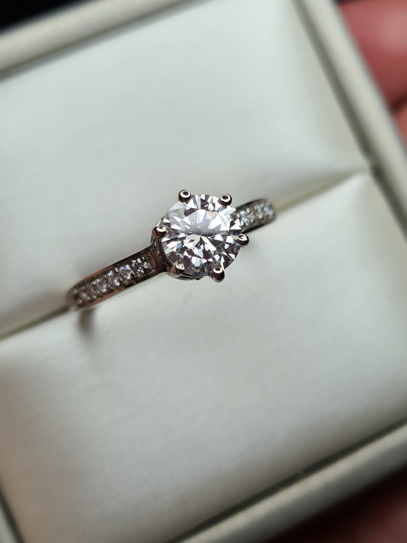 May include: A silver engagement ring with a large, round, clear diamond centre stone and a band of smaller diamonds.