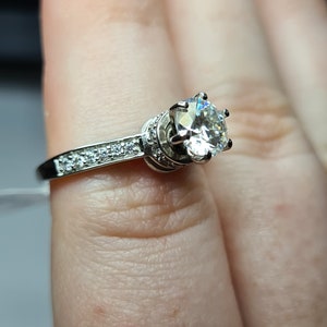 May include: A silver ring with a large, clear, round gemstone surrounded by smaller gemstones. The ring is on a finger.