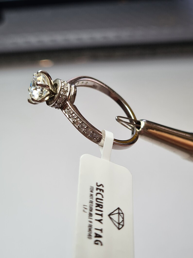 May include: A silver ring with a large, clear diamond set in a prong setting. The ring is surrounded by smaller diamonds. The ring is attached to a security tag that reads "SECURITY TAG ITEM NOT RETURNABLE IF REMOVED L.F.J."
