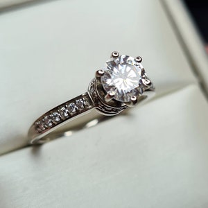 May include: A silver ring featuring a large, clear, round diamond in the centre and smaller diamonds set in a band around the centre stone.