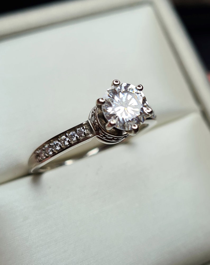 May include: A silver ring featuring a large, clear, round diamond in the centre and smaller diamonds set in a band around the centre stone.