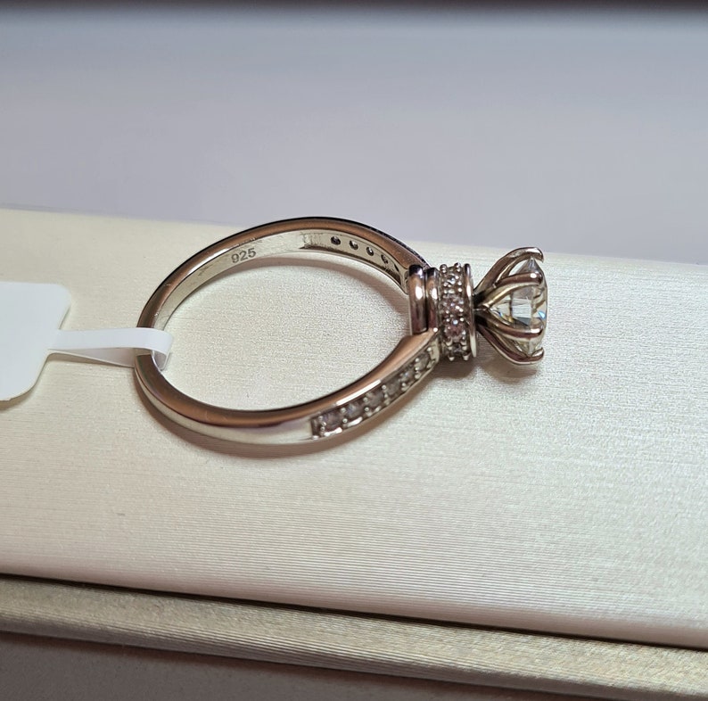 May include: A silver ring with a large, clear centre stone and smaller stones set in a band around the centre stone. The ring is marked "925" on the inside of the band.