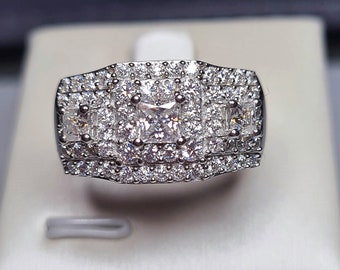 Puede incluir: Un anillo de plata con un gran diamante de forma cuadrada rodeado de diamantes más pequeños. El anillo tiene un diseño de varias capas con varias filas de diamantes más pequeños.