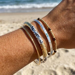May include: A stack of four bracelets including a thin gold-tone wire bangle, a two-tone silver and gold textured cuff with scrollwork, a hammered silver cuff with two small turquoise accents, and a beaded bracelet with gold and dark brown beads.