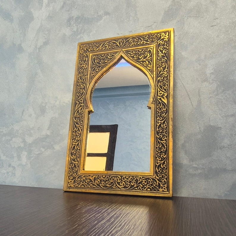 May include: A rectangular gold-framed mirror with an arched top. The frame features intricate black and gold scrollwork. The mirror reflects a glimpse of a room with blue walls and a dark-colored door.