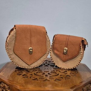 Peut inclure: Deux sacs à bandoulière ronds en cuir marron et toile de jute. Les sacs ont une fermeture à rabat et une couture décorative sur les bords. Les sacs sont de tailles différentes et sont posés sur une surface en bois.