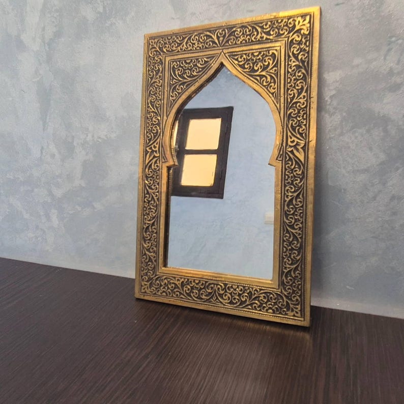 May include: A rectangular mirror with an arched top, featuring a gold frame with intricate black scrollwork. The mirror reflects a window with two yellow panes. The mirror is placed on a dark brown surface against a gray wall.