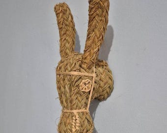 May include: A woven wall hanging in the shape of a rabbit's head and ears. The rabbit is made of natural-colored woven material, with the ears standing upright. The head features a decorative element and is tied with twine.