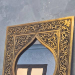 May include: Ornate gold-colored mirror with a decorative, carved frame. The frame features intricate scrollwork and a distinctive arched top. The mirror reflects a glimpse of a window and a blue wall, adding depth to the design.