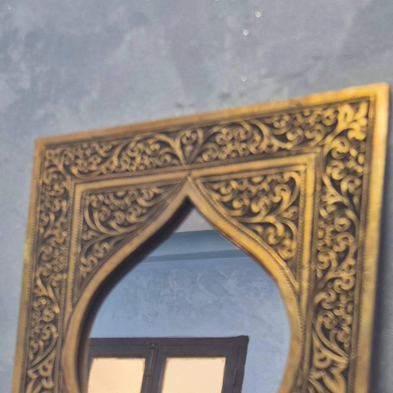 May include: Ornate gold-colored mirror with a decorative, carved frame. The frame features intricate scrollwork and a distinctive arched top. The mirror reflects a glimpse of a window and a blue wall, adding depth to the design.