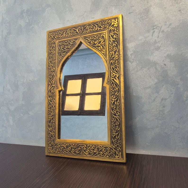 May include: A rectangular mirror with an arched top and ornate gold-colored frame. The frame features intricate black scrollwork. The mirror reflects a window with a dark frame and four gold-colored panes. The mirror is placed on a dark brown surface.