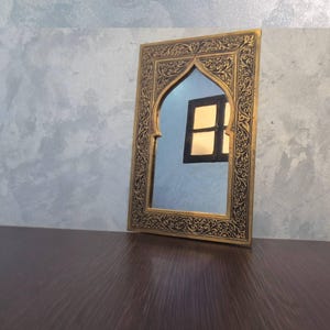 May include: An ornate, rectangular mirror with a gold-colored frame and intricate black detailing. The mirror has an arched top and reflects a window. The mirror is placed on a dark brown surface against a gray wall.