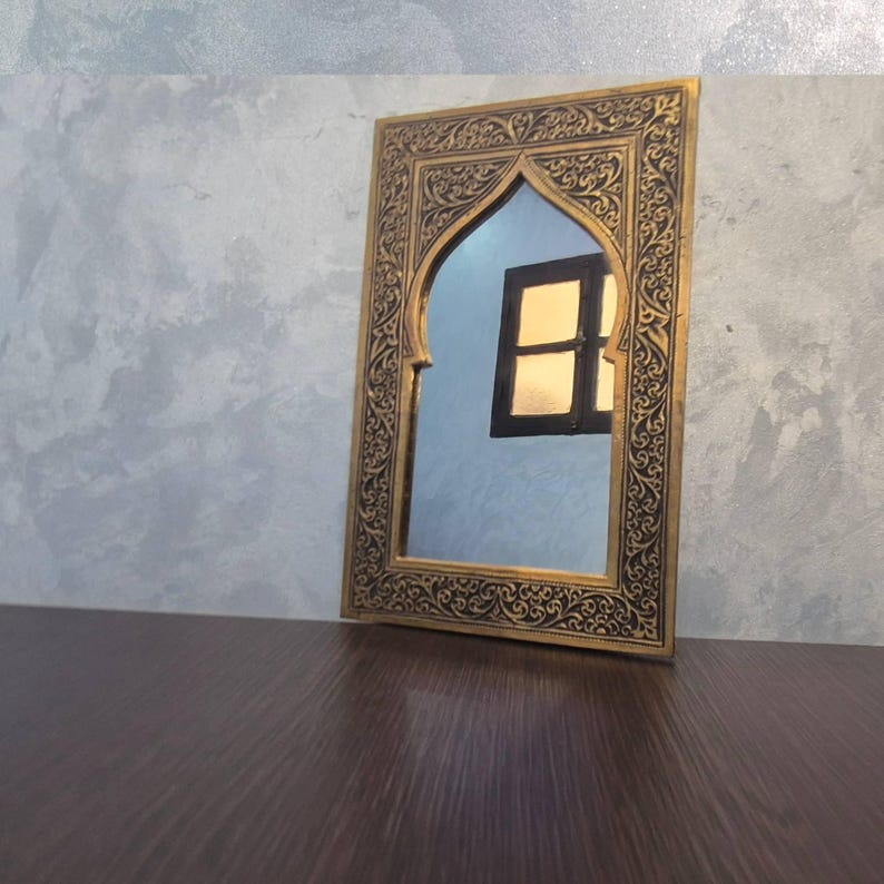May include: An ornate, rectangular mirror with a gold-colored frame and intricate black detailing. The mirror has an arched top and reflects a window. The mirror is placed on a dark brown surface against a gray wall.