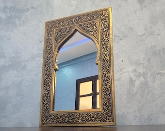 May include: A rectangular mirror with an arched top, framed in gold with detailed black scrollwork. The mirror reflects a room with a blue wall and a dark door, creating a decorative accent piece.