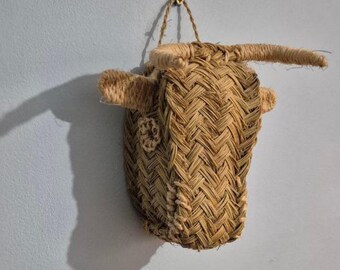 May include: A woven bull head wall hanging. The sculpture is made of natural-colored woven fibers, with horns, ears, and a decorative element. It is suspended from a small hook on a light blue wall. The bull head casts a shadow.