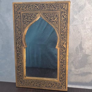 May include: A rectangular, gold-framed mirror with an arched top. The frame features intricate, dark-colored, swirling patterns. The mirror's surface reflects a muted blue hue, creating a decorative piece.