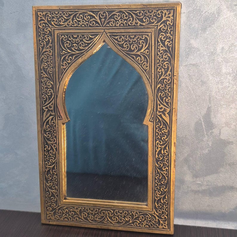 May include: A rectangular, gold-framed mirror with an arched top. The frame features intricate, dark-colored, swirling patterns. The mirror's surface reflects a muted blue hue, creating a decorative piece.