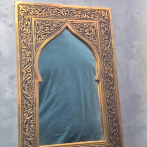May include: Ornate gold-colored mirror with a Moorish arch design. The frame features intricate, dark-colored carvings. The mirror's shape is rectangular with an arched top, reflecting a teal-colored shirt. Decorative wall decor.
