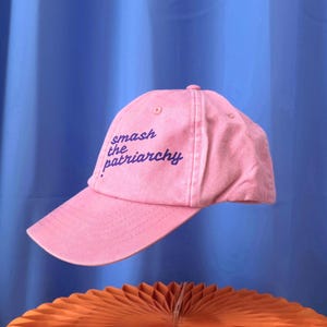 Puede incluir: Una gorra de béisbol de color rosa claro con el texto bordado "smash the patriarchy" en una fuente cursiva de color morado oscuro. La gorra tiene una corona estructurada, una visera curva y pequeños ojales de ventilación en la parte superior.