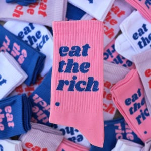 Könnte beinhalten: Eine gerippte, rosafarbene Socke mit dem Text "eat the rich" in dunkelblauer Schrift, die mittig vor einem Hintergrund aus mehreren gefalteten Socken in verschiedenen Farben wie Weiß, Blau und Rosa liegt, die alle denselben Text zeigen.