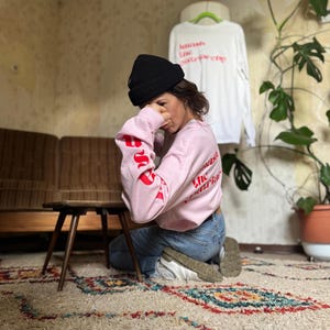 Könnte beinhalten: Eine Person trägt ein pinkfarbenes Langarmshirt mit rotem Text, der "Good Vibes Only" auf dem Ärmel steht. Die Person trägt eine schwarze Mütze und eine blaue Jeans. Die Person sitzt auf einem Teppich vor einer Couch.