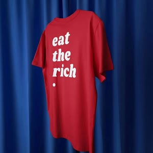 Può includere: Una t-shirt rossa a maniche corte con la scritta bianca "eat the rich" stampata sul davanti. Il capo presenta un classico girocollo e un orlo dritto, creando un netto contrasto tra il tessuto rosso e le lettere bianche stampate sopra.