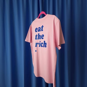 Puede incluir: Una camiseta de manga corta de color rosa claro colgada en una percha de color rosa brillante. La camiseta presenta el texto "eat the rich" impreso en una fuente azul oscuro en la parte delantera, con un pequeño punto azul debajo del texto.