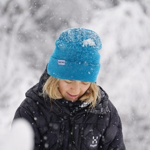 Könnte beinhalten: Eine Person trägt eine schwarze Steppjacke mit einer blauen Strickmütze, auf der "Halu Moi" steht. Die Person steht in einer schneebedeckten Umgebung.