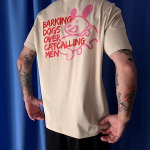 Könnte beinhalten: Ein beiges Kurzarm-T-Shirt mit einer rosa Strichzeichnung eines Hundes auf der Rückseite. Der Text "BARKING DOGS OVER CATCALLING MEN" ist in roter Schrift links neben der Hundegrafik aufgedruckt, die ein Halsbanddetail aufweist.