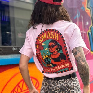 Könnte beinhalten: Hellrosa T-Shirt mit einem Aufdruck auf dem Rücken mit der Aufschrift "SMASH The Patriarchy". Die Grafik zeigt eine Frau mit einem Hammer über einem Globus. Die Person trägt eine rote Kappe und eine Hose mit Leopardenmuster.