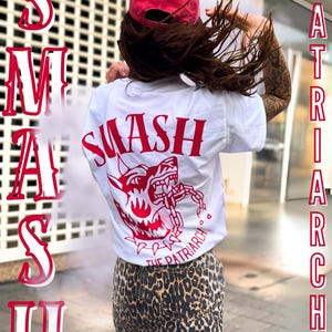 Peut inclure: Une personne porte un t-shirt blanc avec le mot "SMASH" et un dessin rouge d'un chien qui grogne. Le texte "THE PATRIARCHY" est également visible. La personne porte une casquette rouge et une jupe léopard.