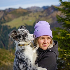 Könnte beinhalten: Eine Frau mit einer lila Mütze mit dem Schriftzug "Maui Now" hält einen schwarz-weißen Border-Collie-Welpen in ihren Armen. Die Frau und der Welpe stehen vor einer Bergkette.