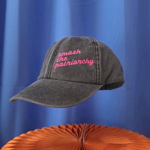 Könnte beinhalten: Eine anthrazitfarbene Baseballkappe mit verwaschener Textur, die auf der Vorderseite den Schriftzug smash the patriarchy in leuchtend pinkfarbener Schreibschrift gestickt hat. Die Kappe hat einen gebogenen Schirm und eine strukturierte Krone.