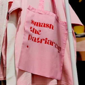 Könnte beinhalten: Eine hellrosa Baumwoll-Tragetasche mit dem roten Schriftzug "smash the patriarchy" auf der Vorderseite, die über einem passenden hellrosa T-Shirt mit kurzen Ärmeln hängt. Die Tasche hat zwei Stoffgriffe und einen kleinen roten Punkt als Grafik.