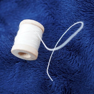 May include: A wooden spool of white thread with a long piece of thread extending from it. The spool is sitting on a blue fuzzy surface.